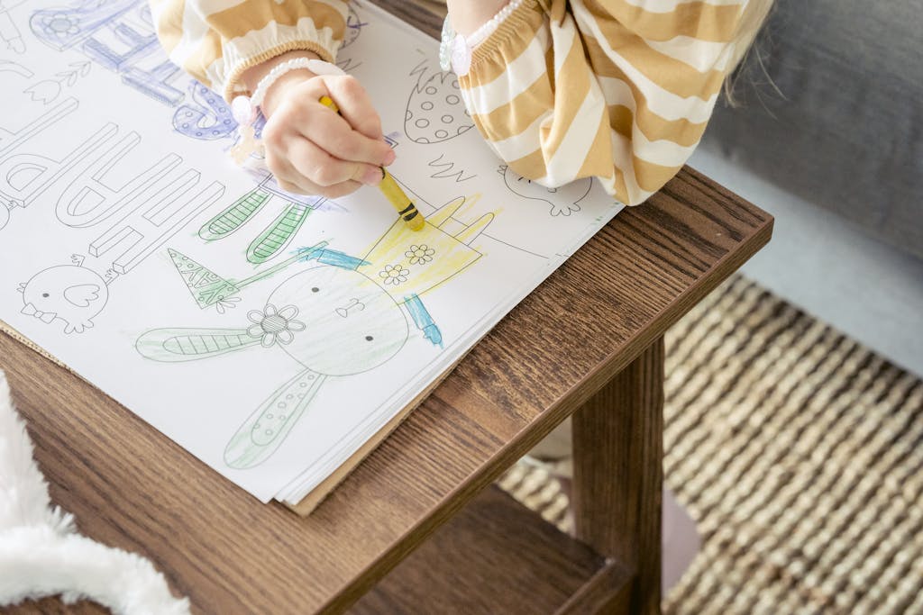 From above of crop anonymous little girl coloring bunny with colorful wax crayon on greeting poster for Easter