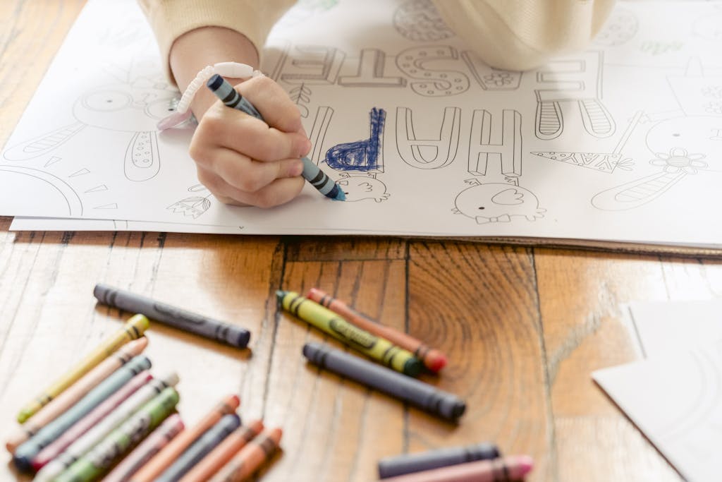 From above of crop anonymous preschool girl drawing with crayons on postcard on floor in daylight